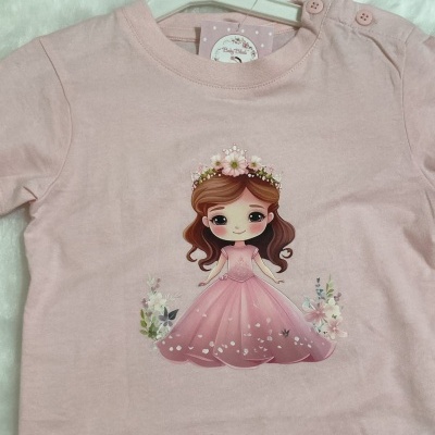 T-shirt de bebé rosa com desenho de menina e flores, pendurada num cabide branco.