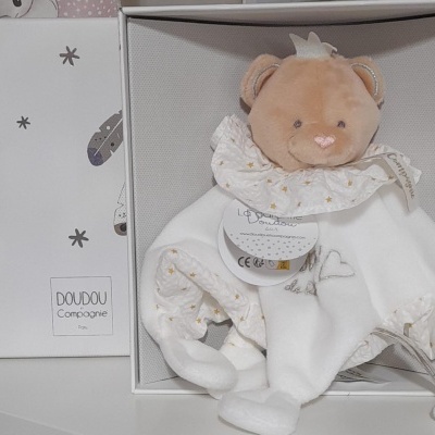 Urso de pelúcia castanho claro com corpo branco em manta, dentro de caixa branca decorada com penas e estrelas