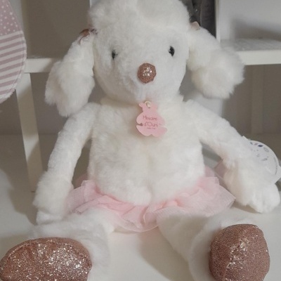 Pelúcia de ovelha branca com focinho e patas cor-de-rosa brilhantes e saia rosa sentada numa prateleira branca.