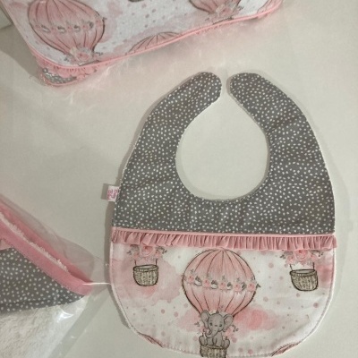 Babete de bebé com padrão balão e elefante, cores rosa, cinzento e nude, com mala e toalha ao fundo.