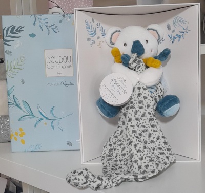 Pelúcia tipo koala branco e azul com lenço estampado em preto e branco em caixa ao lado de embalagem azul clara