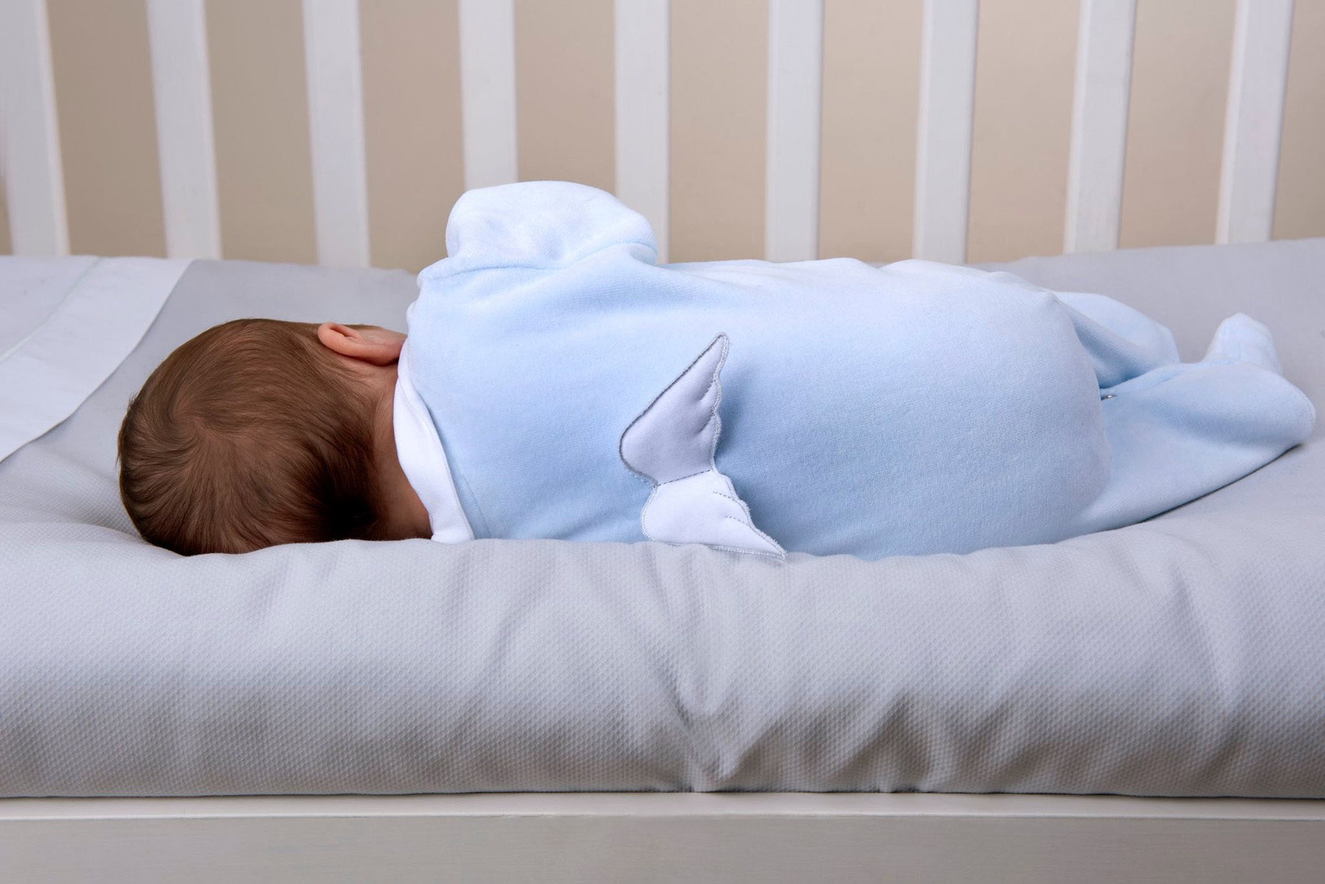 Bebé deitado de barriga para baixo com macacão azul claro com asas brancas numa cama com grades brancas