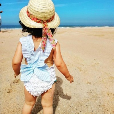 Criança na praia com fato-de-banho branco com laço azul e chapéu de palha com fita florida
