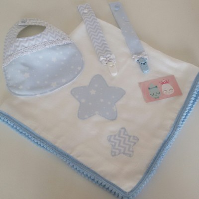 Conjunto de bebé com babete azul, manta branca bordada e fitas prendedoras em tons de azul claro.