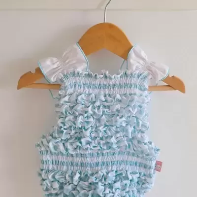 Pelele de bebé com folhos brancos e azuis, gola branca com sanga azul, pendurado num cabide de madeira.