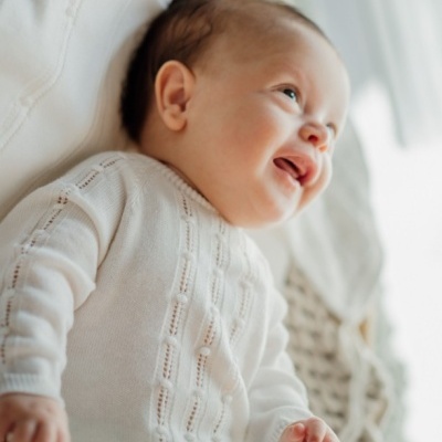 Bebé com macacão branco de malha deitado numa manta trançada bege, sorridente.