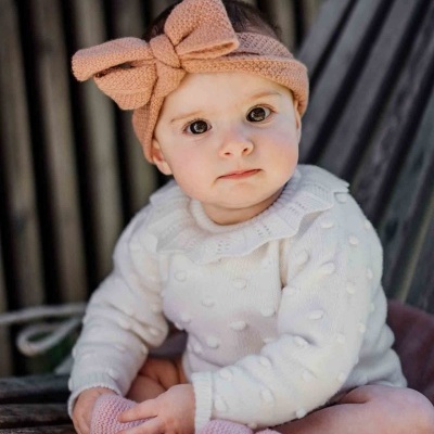 Bebé com camisola branca, faixa de cabeça rosa com laço e meias rosas sentado em superfície de madeira
