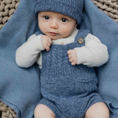 Bebé vestido com macacão e gorro de malha azul cinzento, camisola branca e meias castanhas sobre manta azul num cesto redondo de malha bege