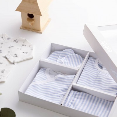 Conjunto de roupa para bebé listrada azul e branca numa caixa branca com babete estampado e casinha de madeira