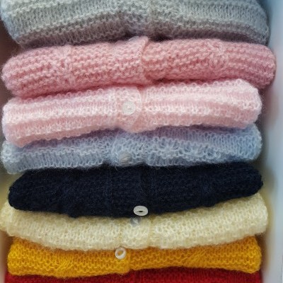 Blusas de malha coloridas empilhadas, cada uma com botão branco, dentro de uma caixa branca