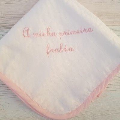 Fralda branca com borda rosa e texto bordado, sobre fundo de madeira clara