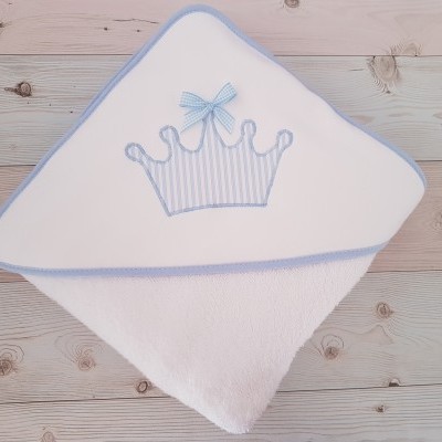 Toalha de bebé branca com capuz azul e aplicação de coroa azul sobre fundo de madeira clara
