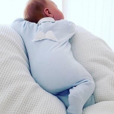 Bebé com babygrow azul claro e asas de anjo sobre almofada branca de malha
