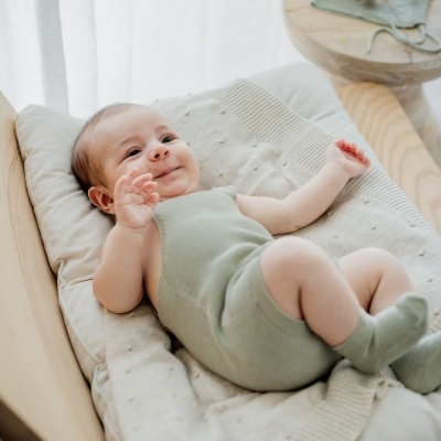 Bebé de macacão verde-oliva deitado em almofada creme numa cadeira de madeira clara, mesa ao lado com roupa verde-oliva