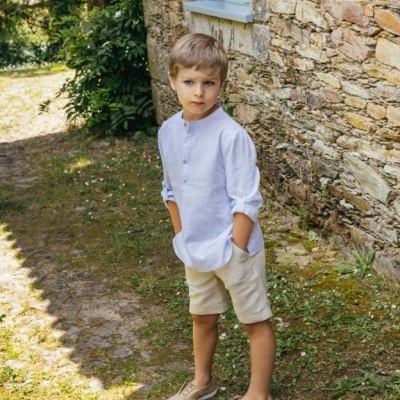 Criança vestindo camisa branca e calções bege ao ar livre junto a parede de pedra