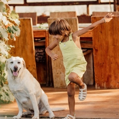 Menina de vestido amarelo claro com sandálias brancas junto a um cão Labrador branco em ambiente rústico exterior.