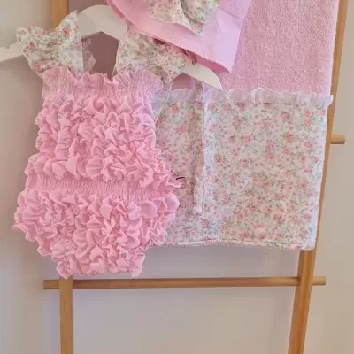 Conjunto infantil rosa com chapéu, body texturado e toalha com faixa floral pendurados numa escada de madeira.