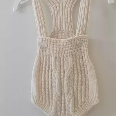 Macacão de bebé em malha creme com alças e botões pendurado num cabide branco