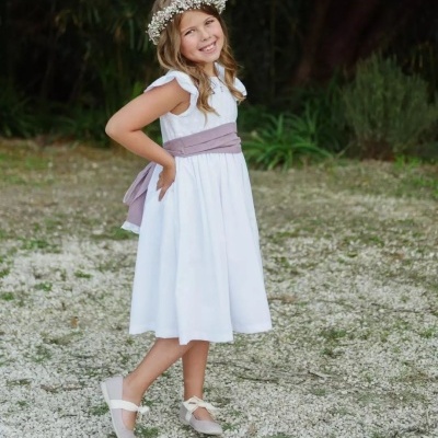 Menina com vestido branco, cinto lilás, sapatilhas claras e coroa de flores num ambiente exterior