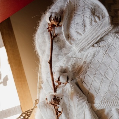 Conjunto de roupa de bebé em malha branca com padrão de losangos, gorro com cordão e peça com botões, manta felpuda e ramo de algodão seco