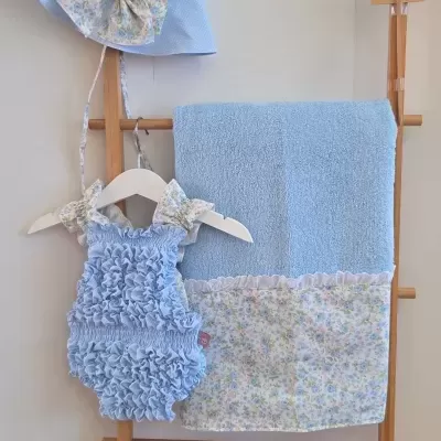 Conjunto infantil azul claro com chapéu, body franzido e toalha com bordado floral pendurados numa estrutura de madeira.