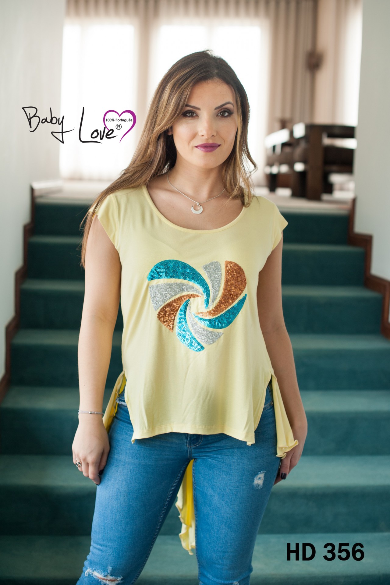 TÚNICA C/ LANTEJOULAS Mulher com t-shirt amarela clara com padrão colorido redondo e calças azuis junto a escadas verdes