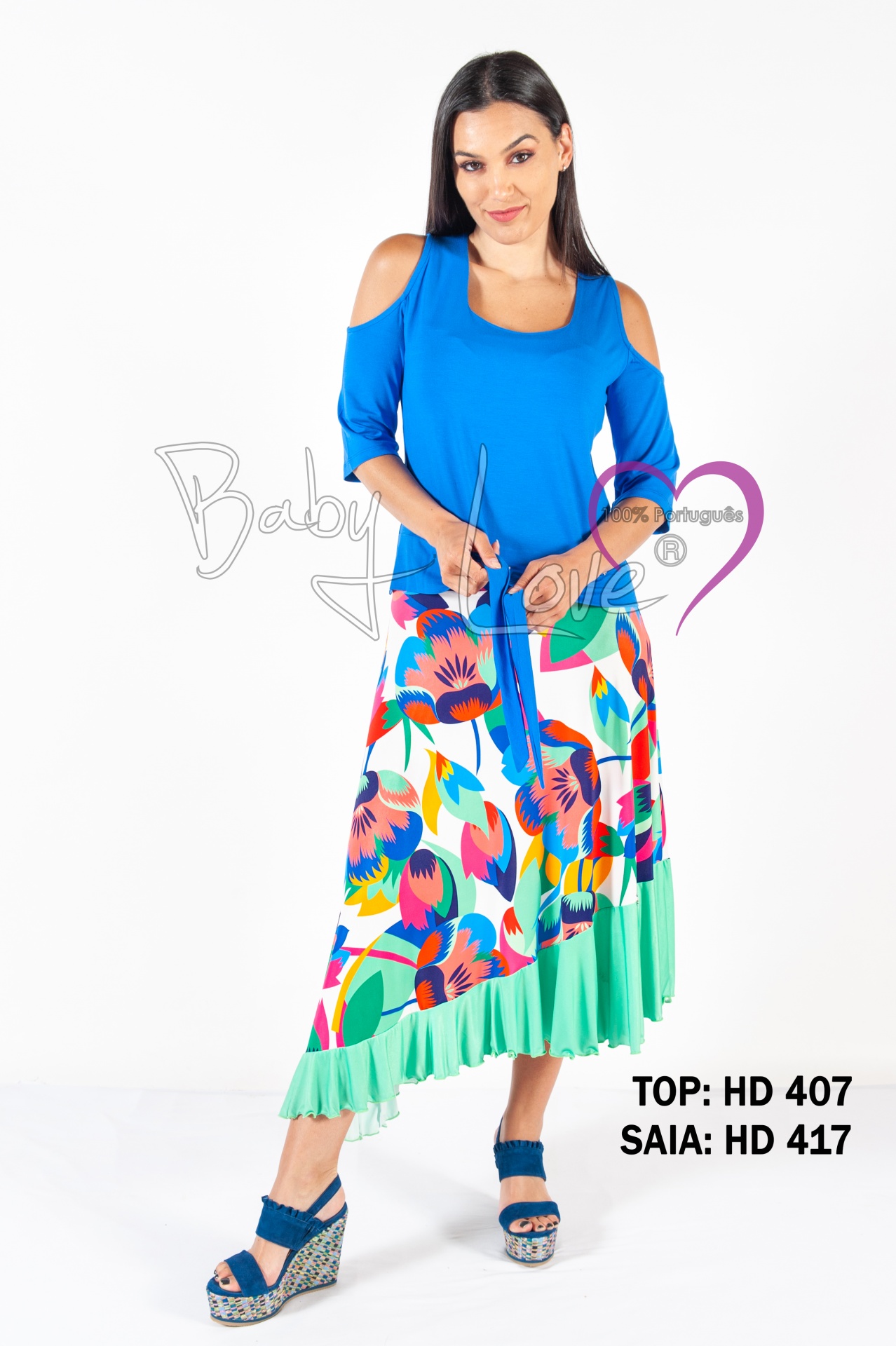 SAIA ASSIMÉTRICA PADRÃO FLORAL Modelo com blusa azul ombros descobertos e saia midi floral colorida com barra verde, texto de referência do produto