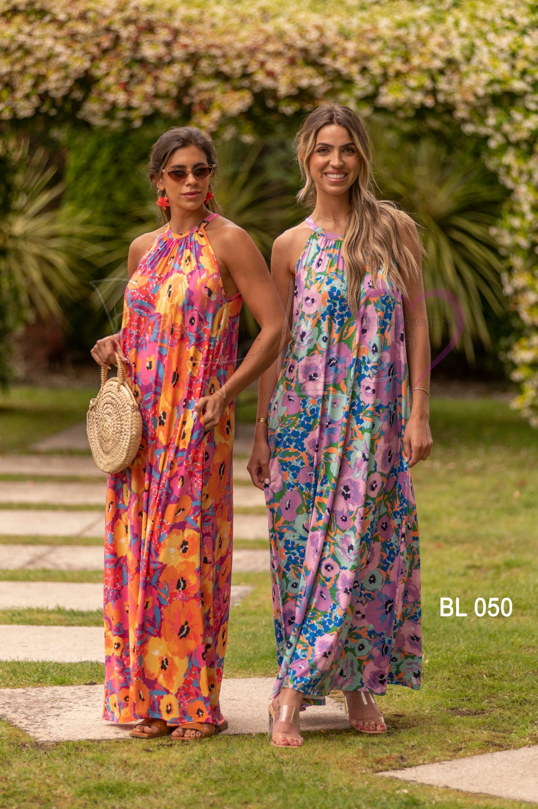 VESTIDO COMPRIDO PADRÃO FLORAL Dois vestidos maxi florais em tecido leve, com padrão colorido e flores grandes, usados por duas mulheres em jardim.