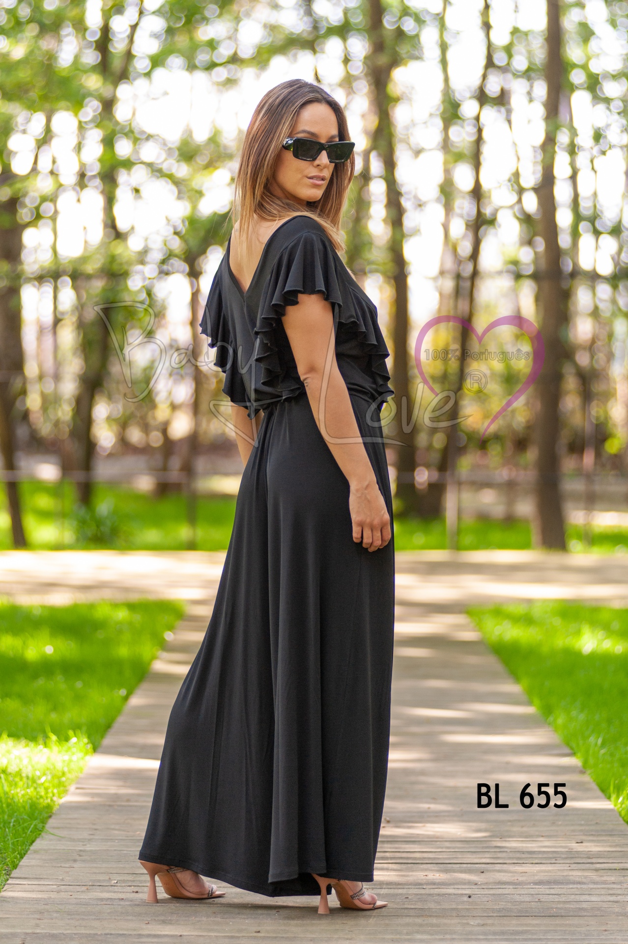 MACACÃO LISO C/ FOLHO Mulher com vestido longo preto de folhos nas costas e mangas, passa por passarela de madeira em parque