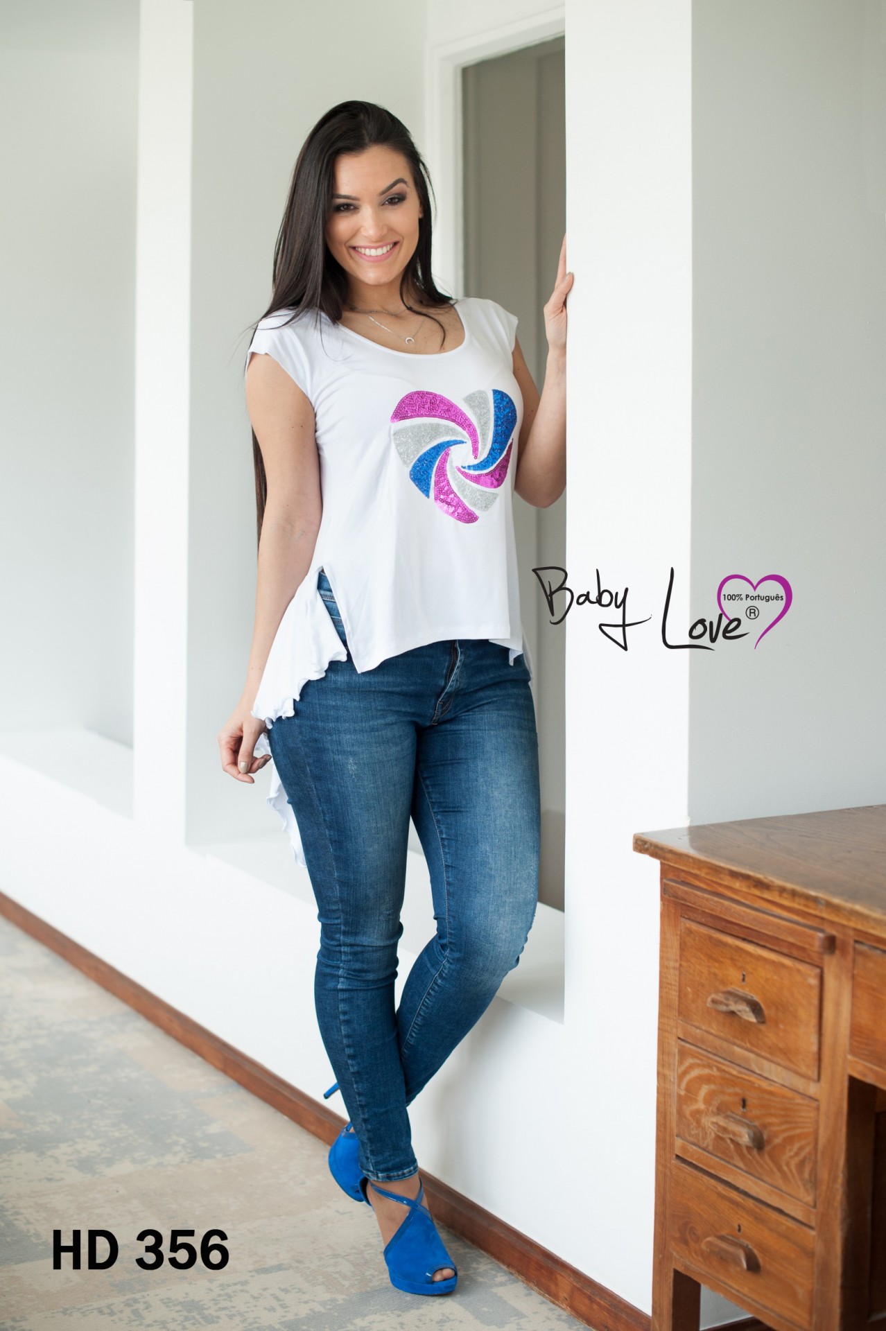 TÚNICA C/ LANTEJOULAS Mulher veste t-shirt branca com estampa colorida, jeans azul e sapatos azuis, parede branca e móvel de madeira ao lado.