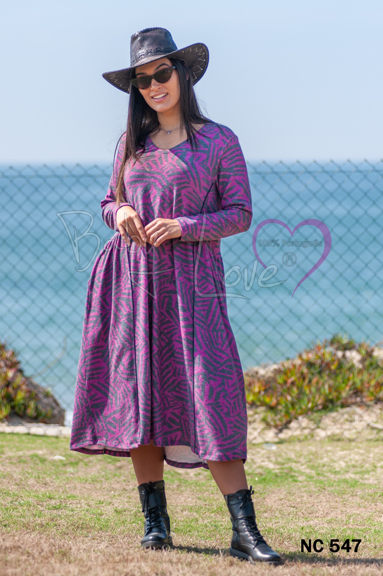 VESTIDO Vestido roxo e rosa com padrão geométrico, chapéu preto e botas junto ao mar