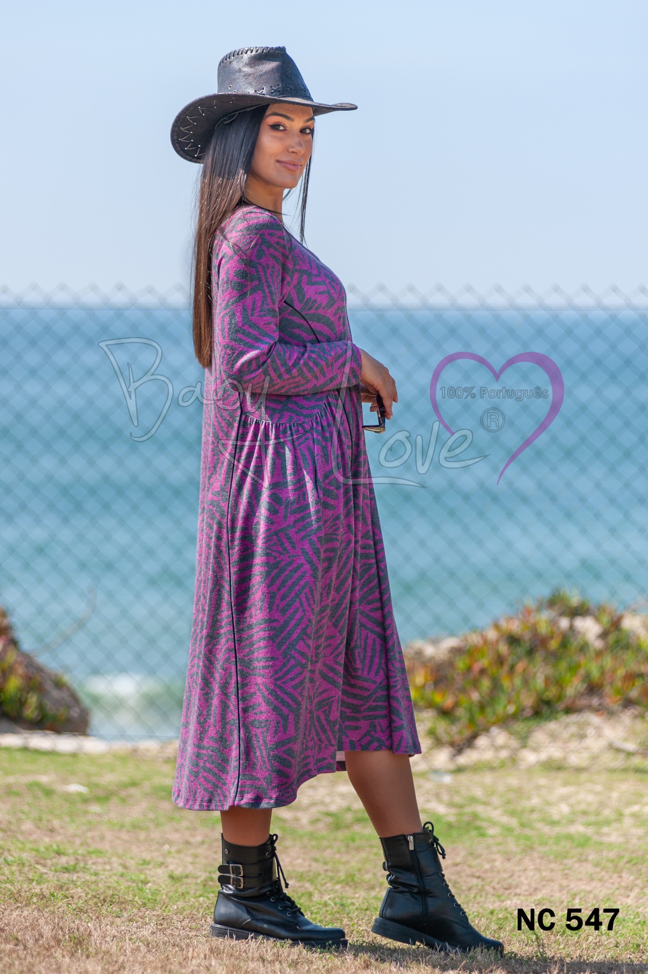 VESTIDO Mulher com vestido rosa e roxo padrão geométrico, botas pretas e chapéu preto numa paisagem ao ar livre com mar ao fundo