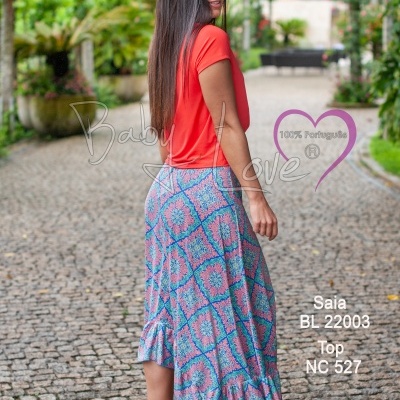 Mulher com saia estampada azul e rosa e blusa laranja em calçada com vegetação