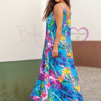 Vestido longo azul com padrão tropical colorido, alças largas, num cenário urbano com parede branca e verde