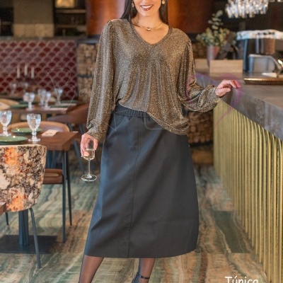 Mulher com túnica dourada e saia preta num restaurante, segurando uma taça.