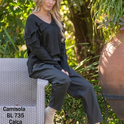 Mulher de camisola preta, calça preta, botas bege e chapéu branco sentada em banco de vime num jardim