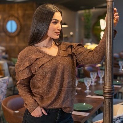 Mulher com túnica castanha brilhante com folhos e calças pretas junto a candeeiro antigo