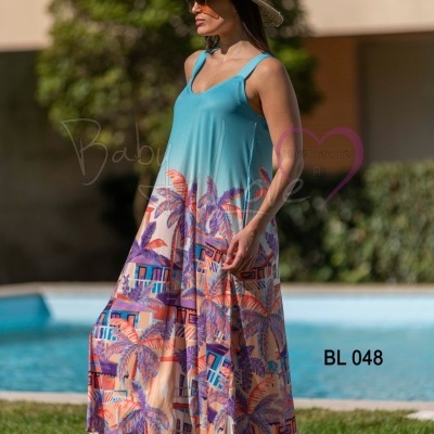Vestido longo azul com estampa colorida de folhagens e casas, usado por mulher com chapéu ao lado de piscina