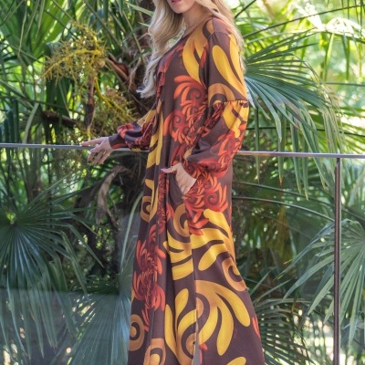 Mulher com vestido comprido castanho com padrão amarelo e vermelho, botas castanhas e boina castanha, em ambiente com folhagem verde.