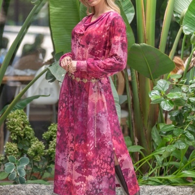 Mulher com vestido longo vermelho e rosa com cinto dourado, botas castanhas, plantas tropicais ao fundo.