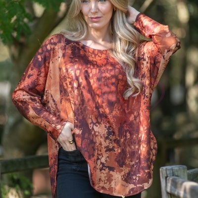 Mulher loira com túnica estampada castanha e vermelha, em ambiente natural com árvores ao fundo