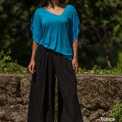 Mulher a usar túnica azul clara e calças largas pretas em ambiente exterior com vegetação.