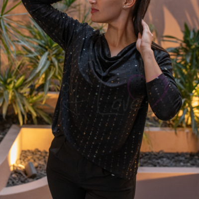 Mulher com blusa preta brilhante e calças pretas em ambiente exterior com plantas