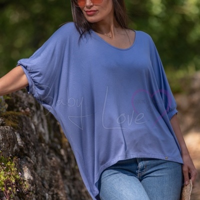 Mulher com túnica azul e calças de ganga encostada a muro de pedra ao ar livre