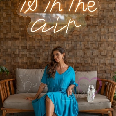 Mulher com vestido azul sentada em sofá sob letreiro de neon 