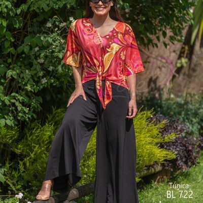 Mulher com túnica colorida e calças pretas ao ar livre junto a plantas.