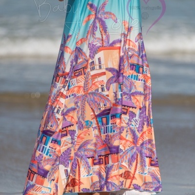 Vestido longo com padrão tropical colorido com palmeiras e casas, em praia com mar ao fundo