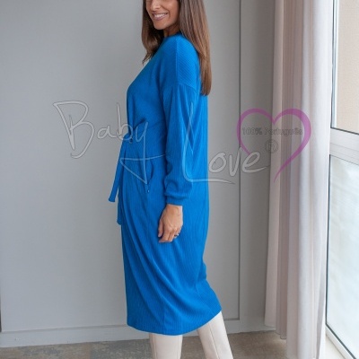 Mulher com vestido azul comprido de malha com cinto e botas brancas, em ambiente interior junto a janela
