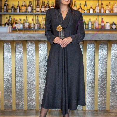 Mulher com vestido cinzento, sapatos castanhos, segurando taça de vidro com limão em bar ao fundo