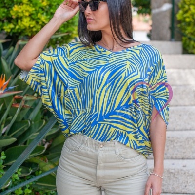 Mulher com t-shirt azul e amarela e calças claras, óculos escuros, ao ar livre